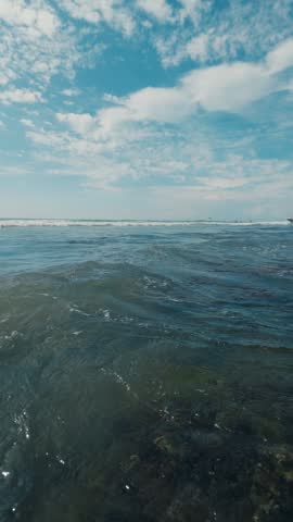 Low-angle ocean waves rolling toward shallow shoreline under blue sky with white