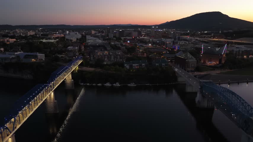 The lit pedestrian Walnut Street Bridge and a second truss bridge cross the dark Tennessee River in Chattanooga at dusk, below Lookout Mountain and an orange afterglow. Aerial drone wide shot