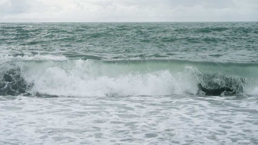 Waves rolling onto a black pebble beach on a grey day in slow motion, Reynisfjara, Vik, Southern Region, Iceland