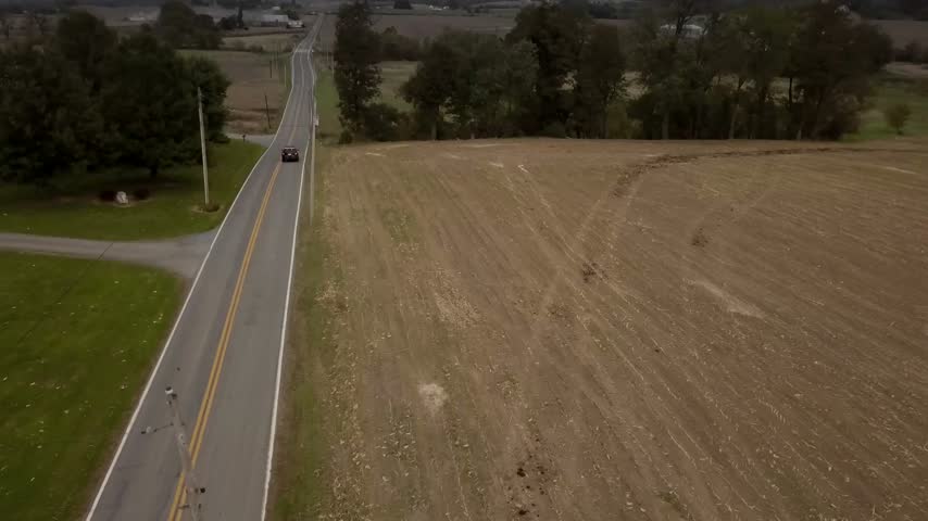 Aerial view of a police SUV cruiser driving along a narrow rural two-lane road flanked by harvested fields and trees in middle America. Establishing tracking drone shot filmed in 2018 in autumn time.