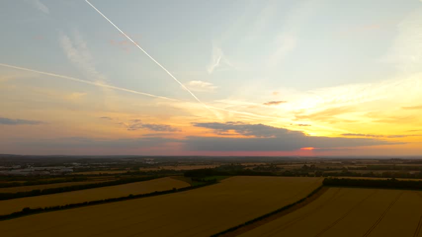 Glowing evening horizon over rolling farmland beneath intersecting contrails and soft clouds. Golden fields with tractor tracks across rural terrain. Sunset countryside, agriculture and peaceful