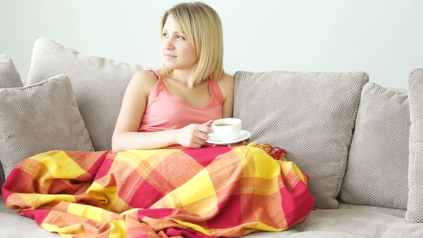 Girl resting on couch with cup of coffee. She covered with blanket
