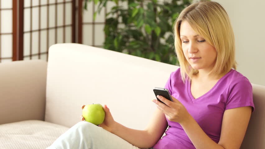 Cute girl with phone sitting on couch and eating an apple
