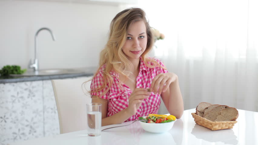 Young woman sitting in kitchen with bowl of salad and glass of water smiling
