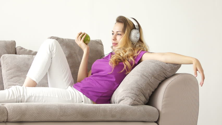 Relaxed young woman lying on sofa eating apple listening to music and smiling at