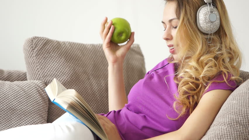 Smiling young woman on sofa reading book listening to music on headphones and