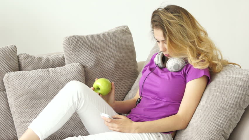 Smiling young  woman relaxing on sofa holding apple in her hand and using mobile