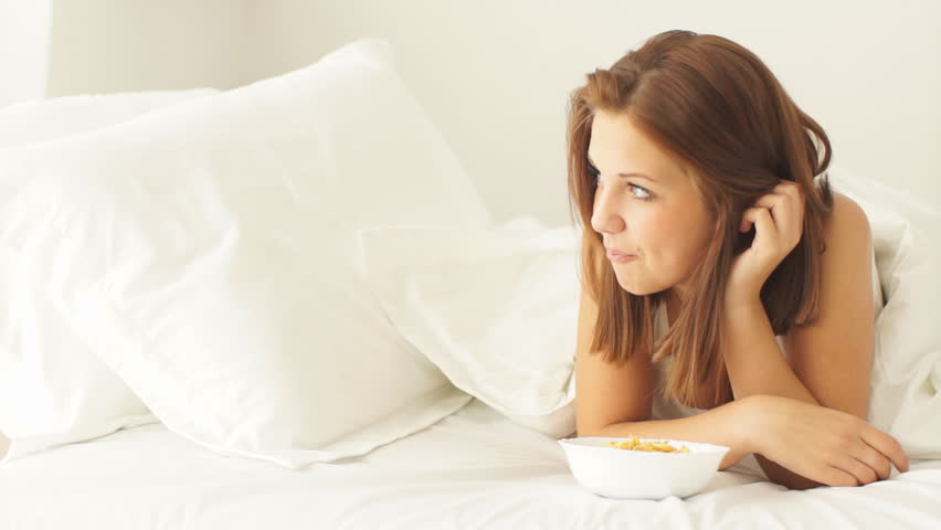 Smiling girl lying in bed eating corn flakes and laughing at camera