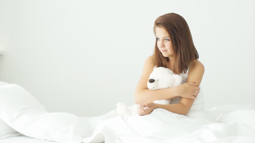 Young woman sitting on bed playing with teddy bear and smiling at camera