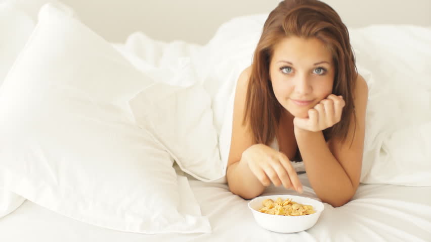 Young woman lying in bed eating corn flakes and smiling at camera