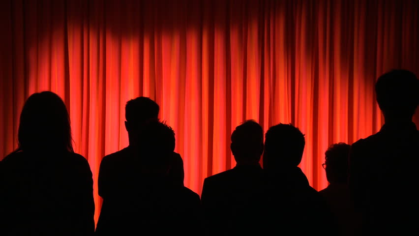 Silhouetted theater audience waits in front of red curtains.  Curtains open and