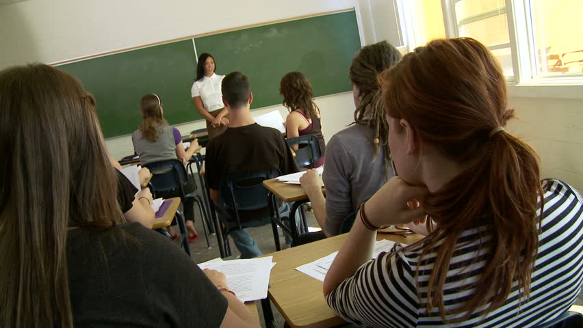 Camera at back of a high school classroom with woman teaching at the front of a