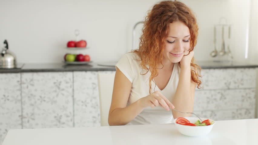Smiling young woman sitting at kitchen table and eating vegetable salad
