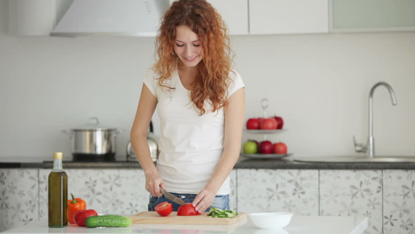 Smiling young woman standing in kitchen slicing tomatoes
