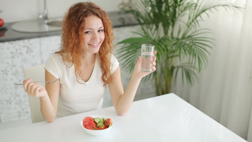 Smiling young woman sitting at kitchen table holding glass of water and eating