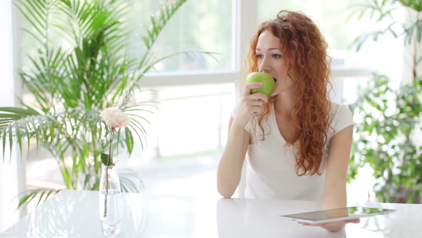 Young woman sitting at table using touchpad eating apple and smiling at camera
