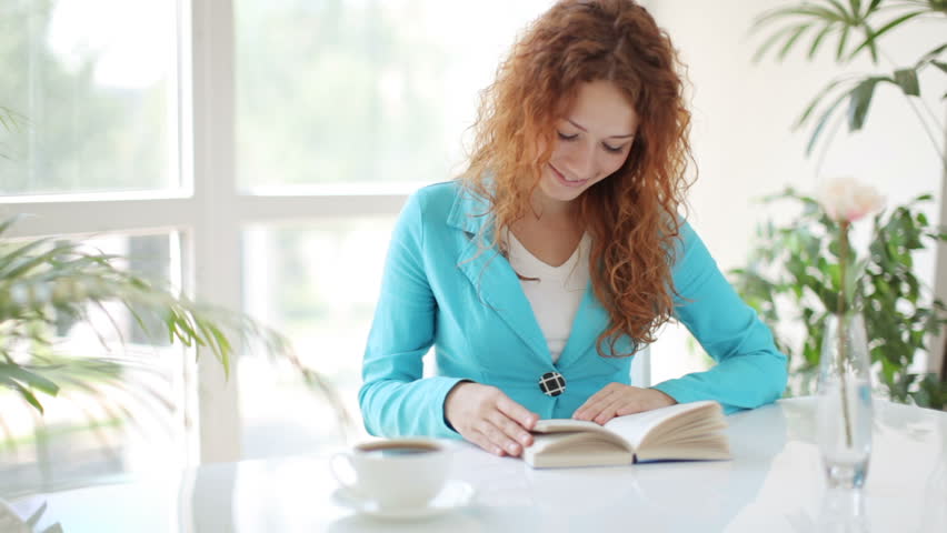 Young woman sitting at table reading book and smiling at camera
