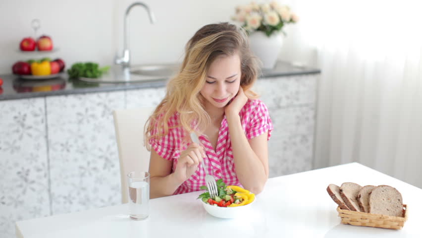 Cute blond girl sitting at table in kitchen and eating salad
