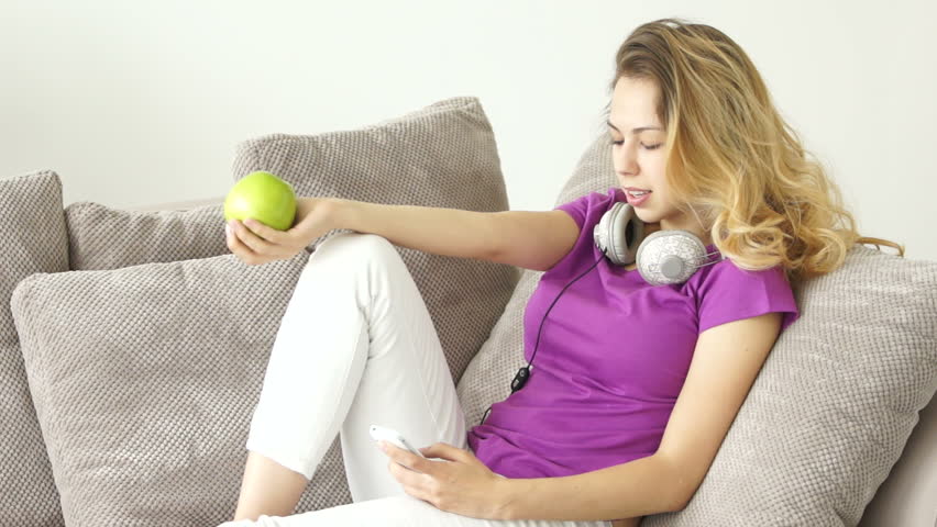 Happy young woman on sofa biting green apple and using mobile phone
