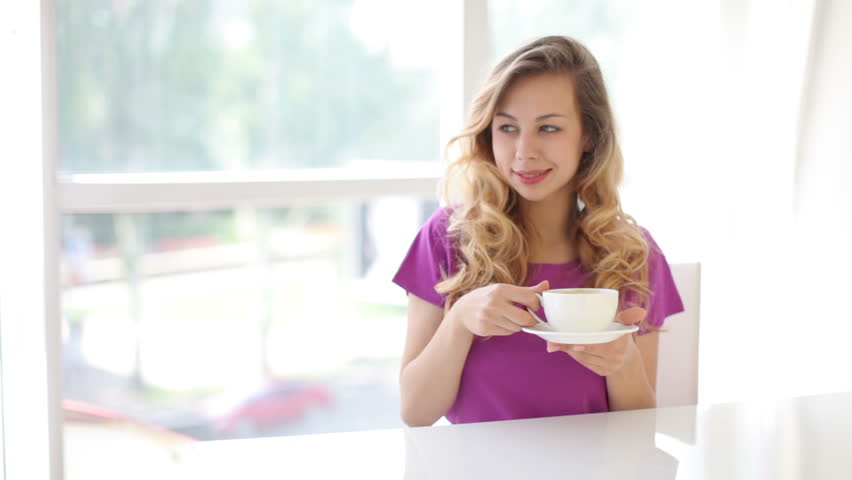 Young woman sitting at table drinking coffee and looking at camera with smile
