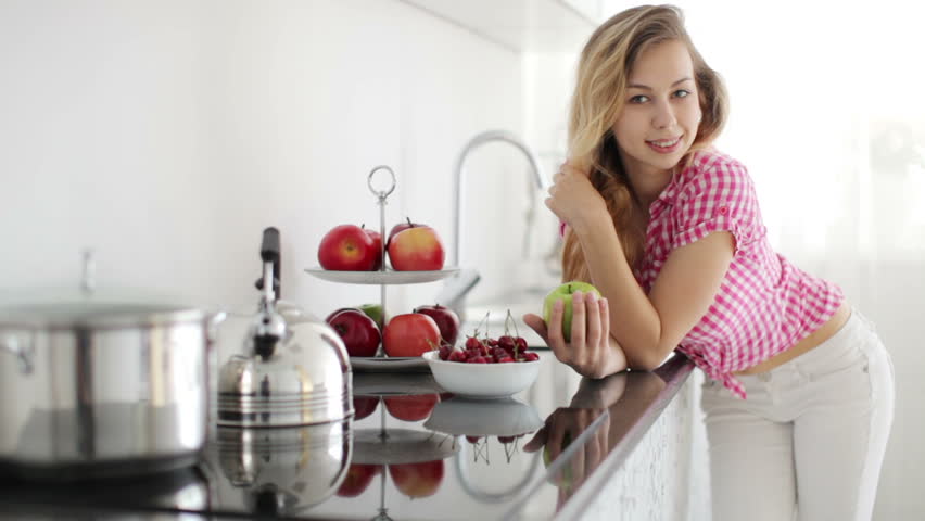 Pretty girl in kitchen holding green apple and looking at camera with smile
