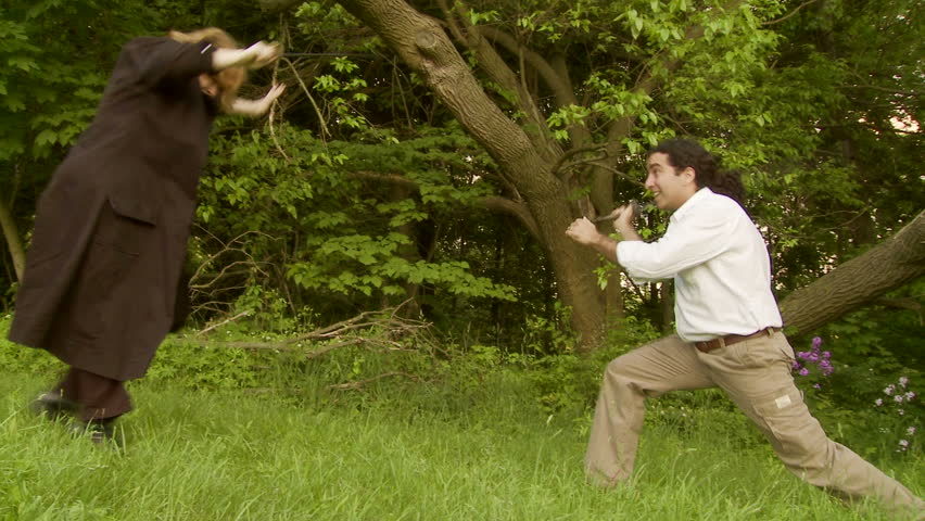 Couple duel with fencing swords on grassland in front of woods.