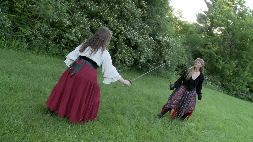 Two women in medieval-style costumes fight with swords on a grassy field.