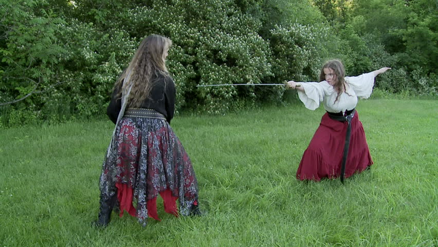 Two women in medieval-style costumes fight with swords on a grassy field.  Slow