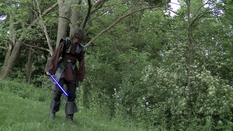 Two men duel with futuristic glowing blue laser swords.  Real time with normal shutter speed on grassland in front of woods.  Hand held camera.