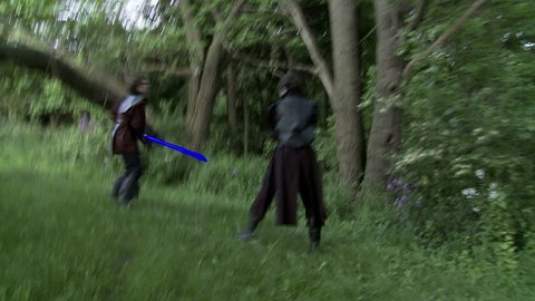 Two men duel with futuristic glowing blue laser swords.  Fighting on grassland in front of woods.  Hand held camera.  Long shutter speed gives strange movement and large light trails from the lasers.