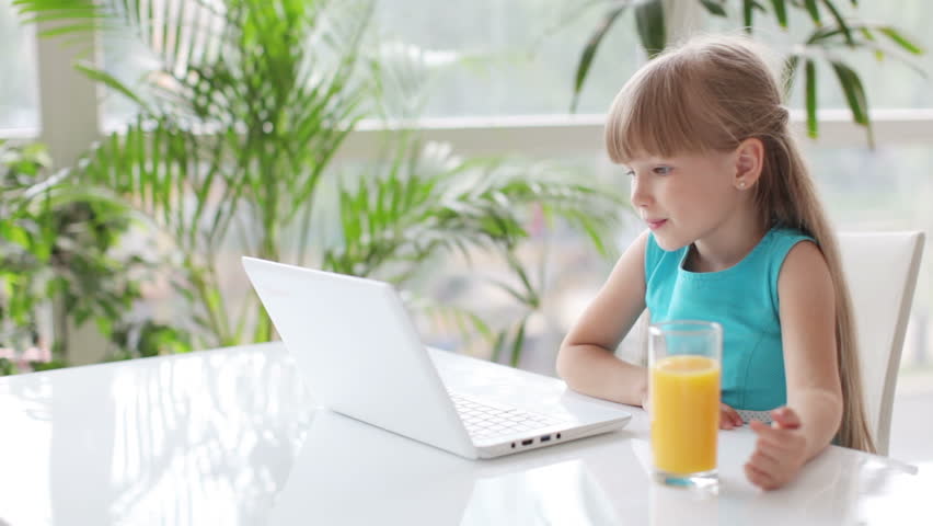 Pretty little girl sitting at table drinking juice using laptop and smiling
