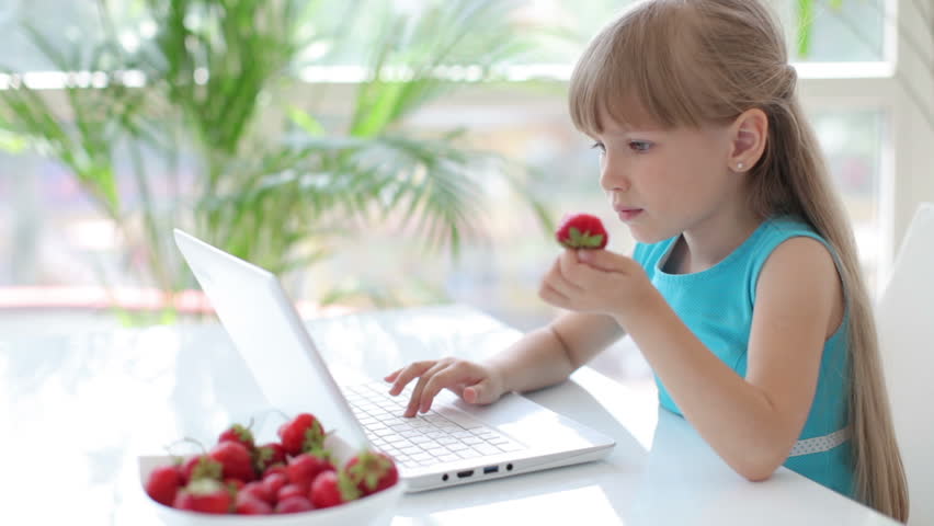 Little girl sitting at table eating strawberries using laptop smiling and