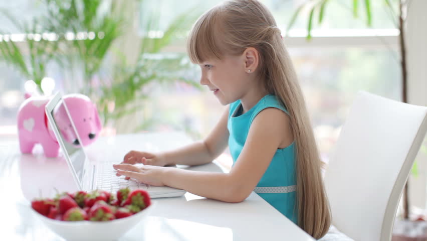 Funny little girl sitting at table using laptop and looking at camera smiling
