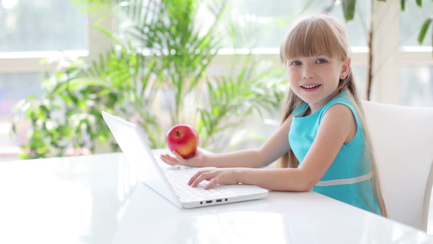 Funny little girl sitting at table holding red apple in her hand using laptop
