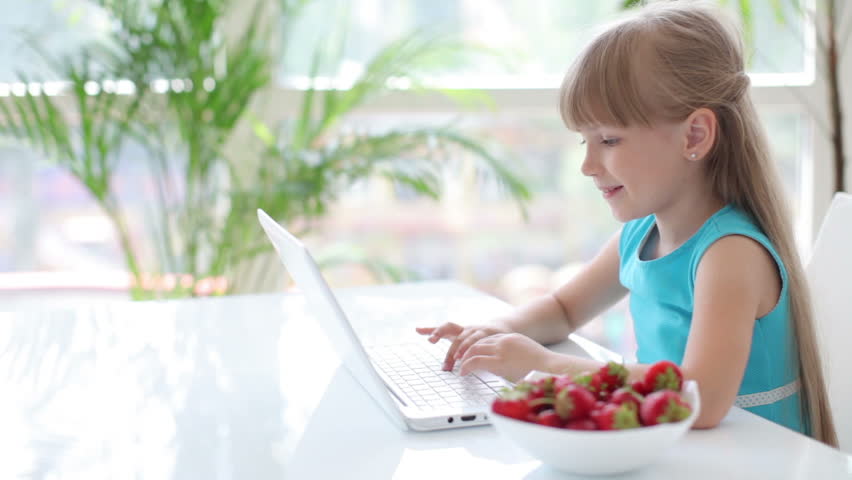 Funny little girl sitting at table using laptop eating strawberries and smiling