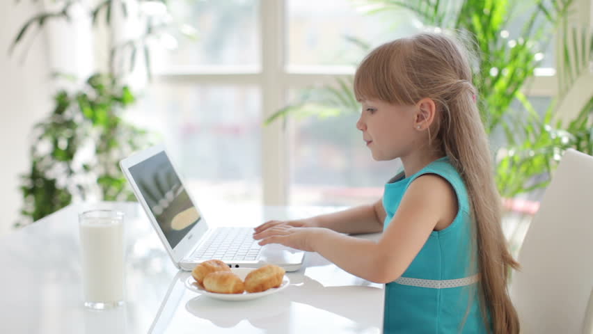 Funny little girl using laptop and eating cakes and looking at camera smiling
