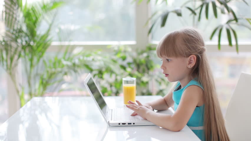 Little girl sitting at table using laptop and looking at camera with smile
