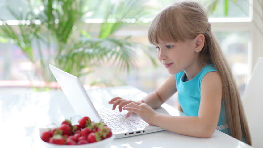 Funny little girl sitting at table with laptop eating strawberries and making