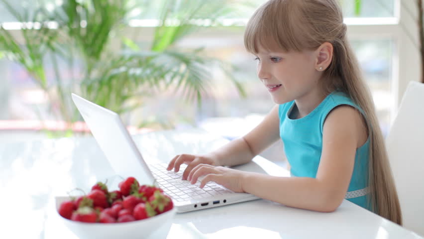 Pretty little girl sitting at table eating strawberries using laptop and smiling