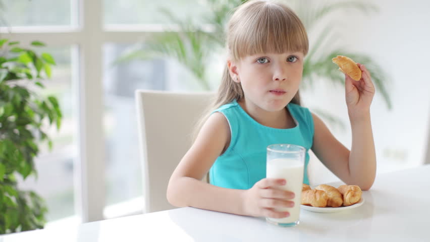 Funny little girl sitting at table drinking milk and eating cakes and smiling at