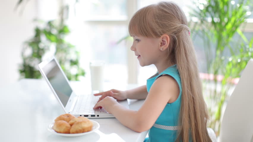 Funny little girl sitting at table eating cake and using laptop
