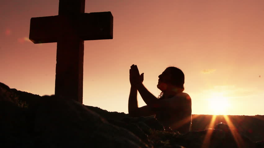 Silhouette of man praying under the cross at sunset/sunsrise