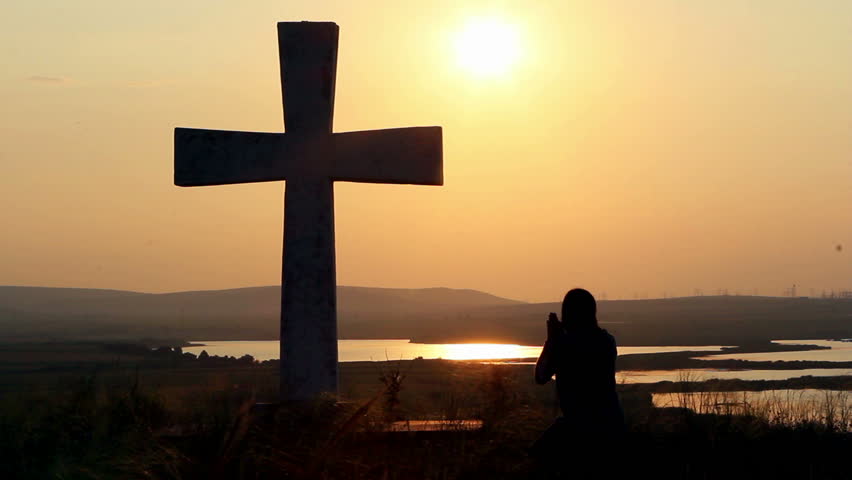 Silhouette of man praying under the cross at sunset/sunsrise
