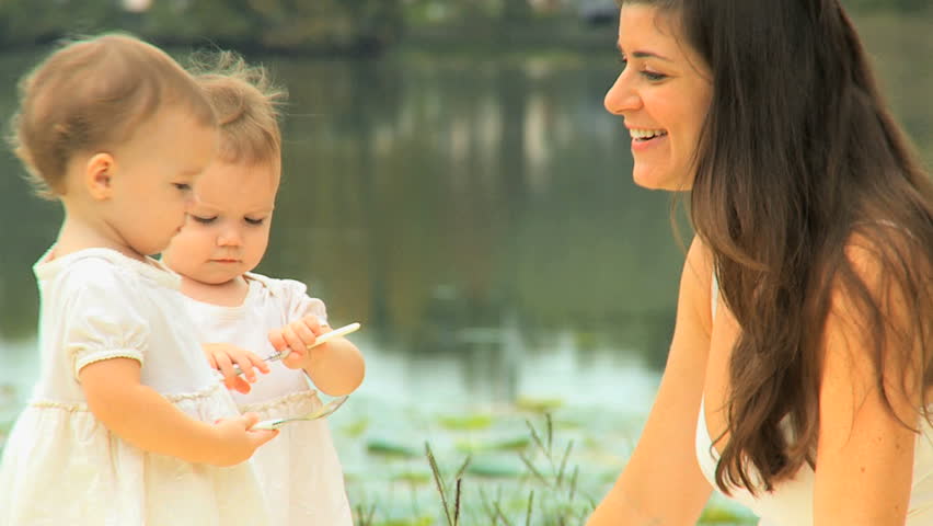 Happy young mom with twin blonde toddler girls having fun on grass by lake in park close up