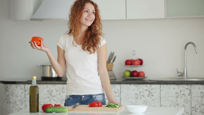 Pretty young woman standing in kitchen cutting vegetables and smiling at camera
