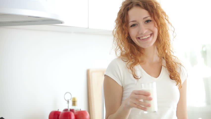 Pretty young woman standing in kitchen drinking milk from glass and smiling at