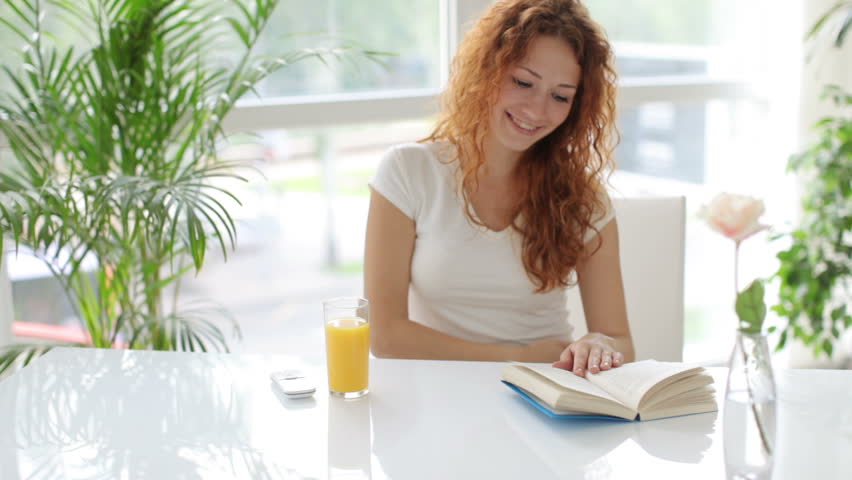 Pretty young woman sitting at table and reading book with smile on her face
