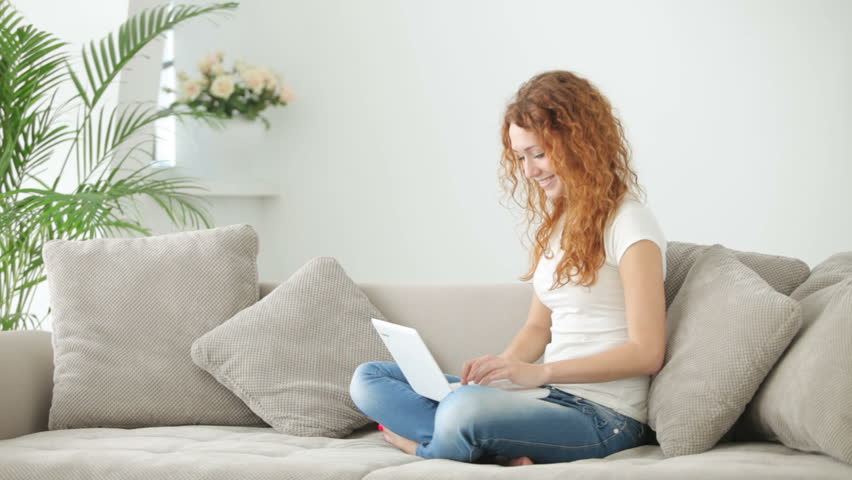 Pretty young woman sitting on sofa using laptop and smiling at camera
