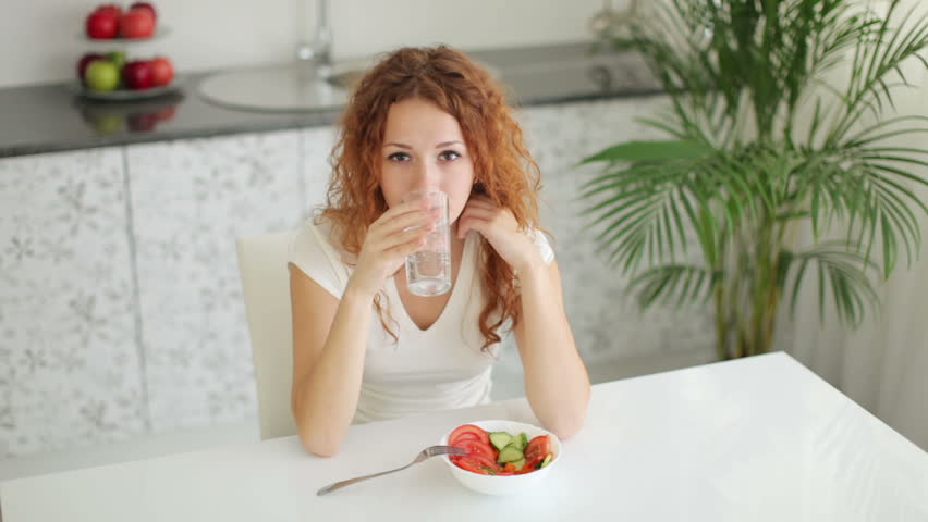Young woman sitting at kitchen table drinking water and smiling at camera
