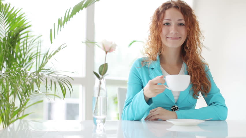 Pretty young woman drinking coffee at table and looking at camera with smile
